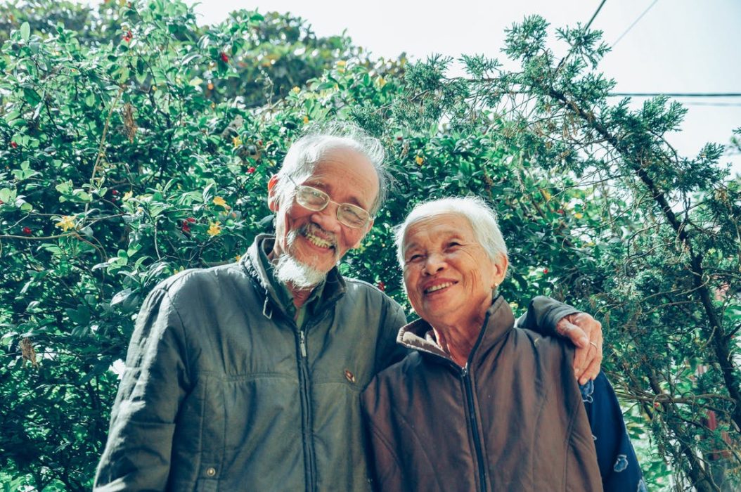 Elderly smiling couple in front of greenery outside.