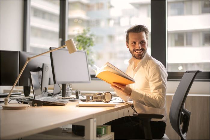 Man holds book and sits at desk with raised desktop monitor.