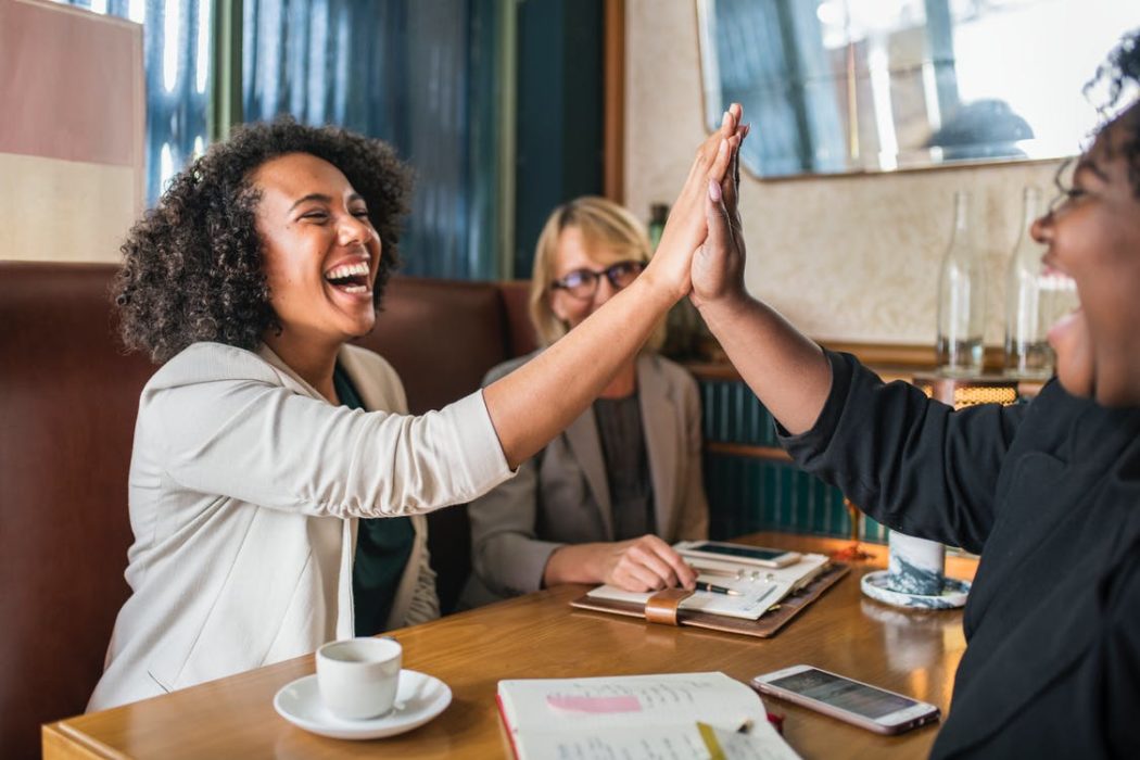 Three women sitting at cafe booth. Two of the women high five.